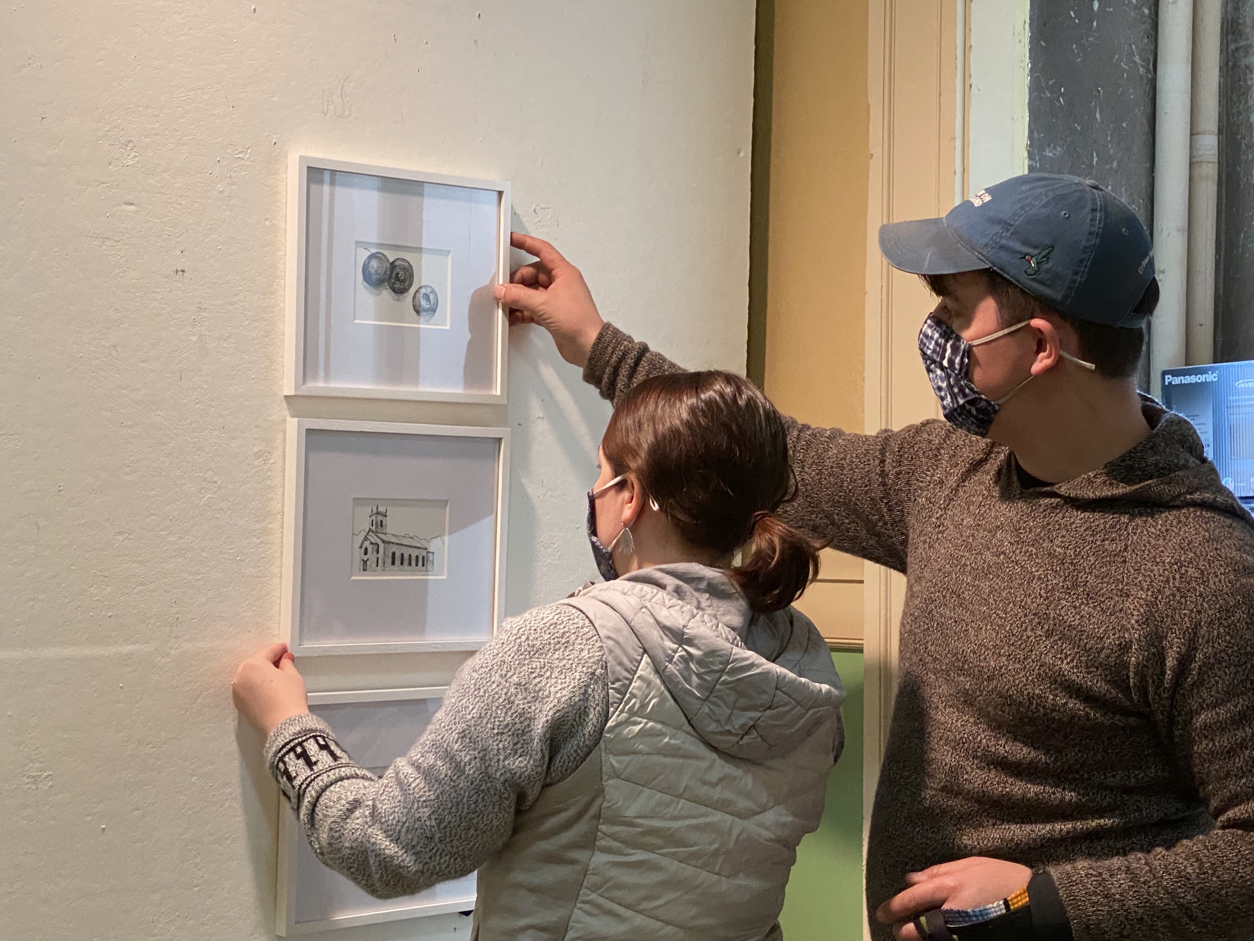 Artist Amy Hook Therrien in white knobby sweater is assisted by partner Alex wearing baseball cap to hang a series of 3 smaller pen & ink drawings in a verticle line on display panel at LNT